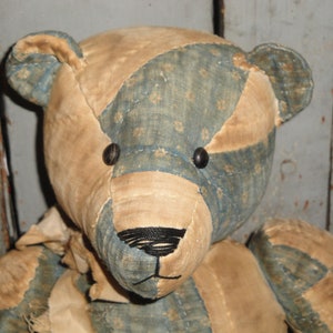 May include: A patchwork teddy bear with blue and tan fabric. The bear has black button eyes and a black embroidered nose.