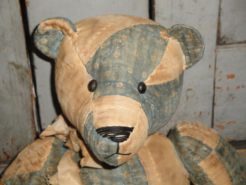 May include: A patchwork teddy bear with blue and tan fabric. The bear has black button eyes and a black embroidered nose.