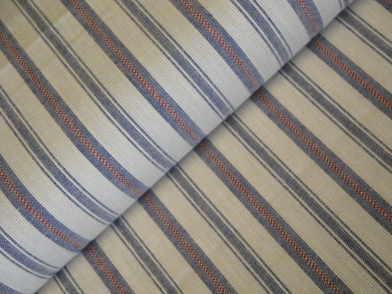 May include: Cream-coloured fabric featuring a repeating pattern of blue and red stripes. The stripes run diagonally across the fabric, creating a visually interesting texture. The fabric appears to be folded over itself.