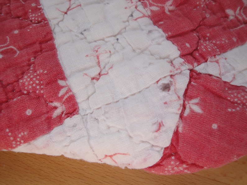 Op de afbeelding: Een doorgestikt stuk stof met afwisselende verticale strepen in rood en wit. De rode delen zijn voorzien van kleine witte bloemmotieven, terwijl de witte delen effen zijn. De stof heeft een zichtbare doorgestikte textuur.