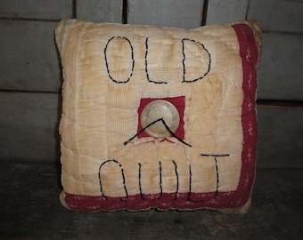 May include: A white pillow with red trim and black embroidery that reads "OLD QUILT". A silver button is sewn in the center of the pillow.