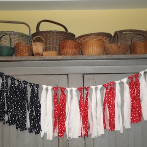 May include: A patriotic-themed garland with alternating sections of black fabric with white stars, solid white, and red fabric with white polka dots. Woven baskets of various sizes and shapes are displayed on a shelf above the garland.