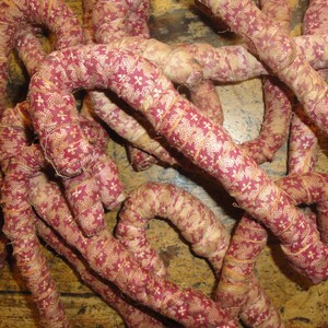 K&ouml;nnte beinhalten: Eine Ansammlung mehrerer stoffbezogener Zuckerstangenformen. Jedes St&uuml;ck ist mit einem dunkelroten und cremefarbenen Textil umwickelt, das ein sich wiederholendes kleines Blumen- und Kreuzmuster aufweist. Die flexiblen, l&auml;nglichen Formen sind gebogen.