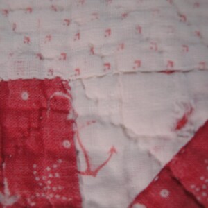 Op de afbeelding: Een doorgestikte stof met een patchworkontwerp met afwisselende secties van witte stof met kleine rode ankermotieven en effen rode stof met witte stippenpatronen, waarbij de stiksels door het hele materiaal heen zichtbaar zijn.