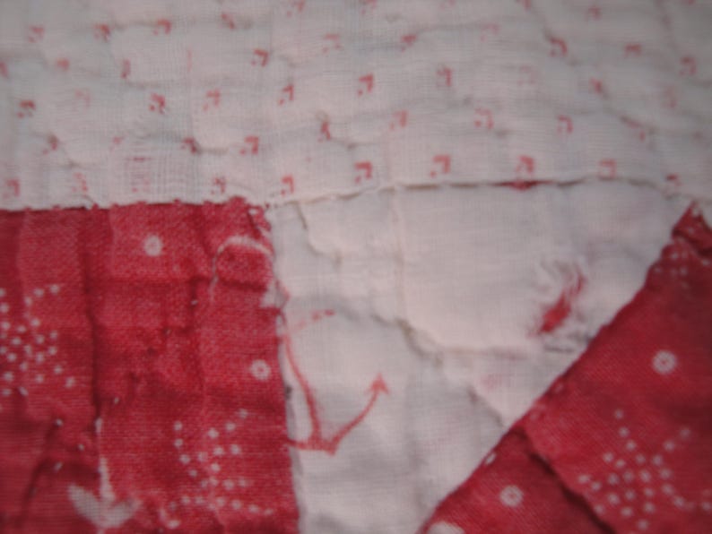 Op de afbeelding: Een doorgestikte stof met een patchworkontwerp met afwisselende secties van witte stof met kleine rode ankermotieven en effen rode stof met witte stippenpatronen, waarbij de stiksels door het hele materiaal heen zichtbaar zijn.