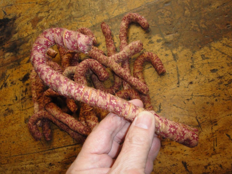 K&ouml;nnte beinhalten: Eine Sammlung von stoffbezogenen Zuckerstangenformen mit einem dunkelroten und cremefarbenen Blumenmuster. Jede Stange besteht aus gewickeltem Textilmaterial und zeigt ein gebogenes Oberteil sowie einen geraden Schaft in einer Gruppe.
