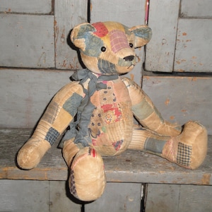 May include: A patchwork teddy bear constructed from various fabric scraps in shades of tan, blue, red, and plaid. The bear features dark button eyes, a stitched nose, and a frayed dark gray ribbon tied around its neck, with visible seams throughout.
