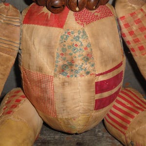 May include: A patchwork doll with a brown wooden head and a bell necklace. The doll is made of various fabrics in shades of red, white, blue, and green.