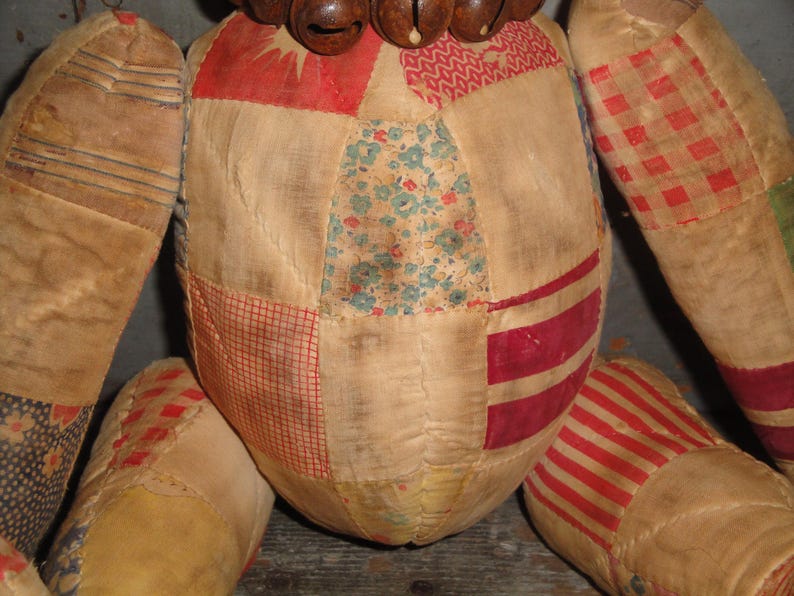 May include: A patchwork doll with a brown wooden head and a bell necklace. The doll is made of various fabrics in shades of red, white, blue, and green.