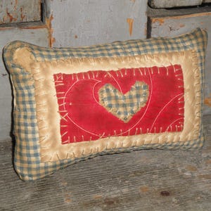 May include: A rectangular pillow with a red heart design. The pillow features a red center with a plaid heart, surrounded by a tan border and a blue and tan checkered fabric. The edges are stitched.