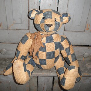 May include: A patchwork teddy bear with a checkered pattern in blue and tan. The bear is sitting on a wooden surface.