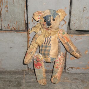 May include: A vintage teddy bear made from patchwork fabric. The bear has a tan body with patches of blue, orange, and plaid fabric. It has black button eyes and a stitched nose. The bear is in a sitting position.