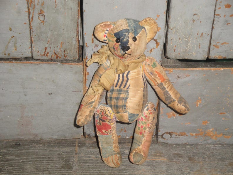 May include: A vintage teddy bear made from patchwork fabric. The bear has a tan body with patches of blue, orange, and plaid fabric. It has black button eyes and a stitched nose. The bear is in a sitting position.