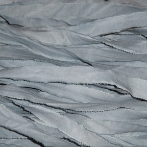 May include: Close-up of a textured fabric in shades of gray-blue. The fabric is composed of layered, wavy strips with frayed edges, creating a tactile and visually interesting surface. The overall effect is soft and flowing.