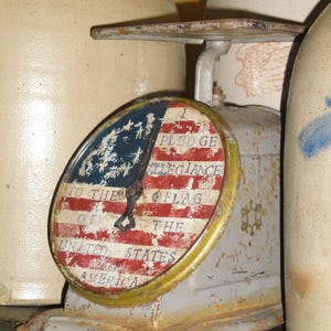 May include: An antique scale with a rusty metal base and a round, white, red, and blue dial that reads "I Pledge Allegiance to the Flag of the United States of America".