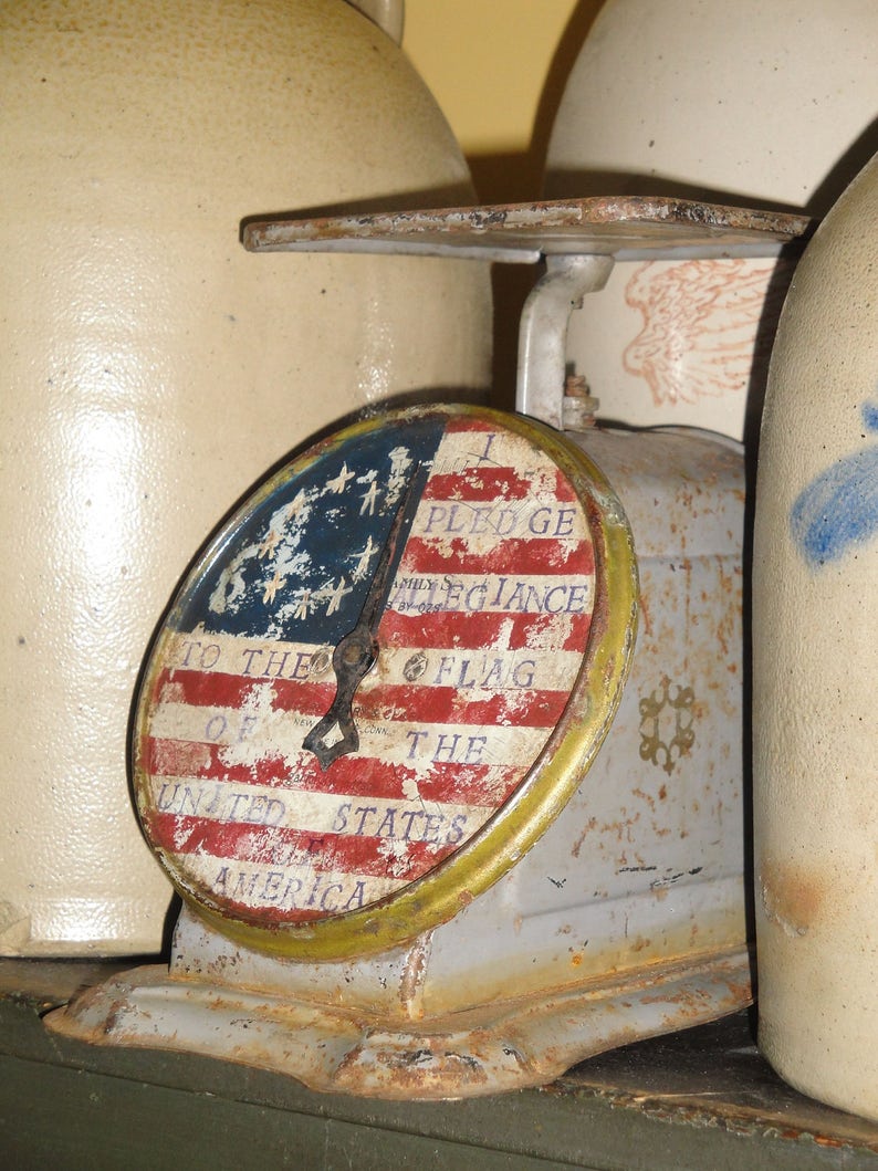 May include: An antique scale with a rusty metal base and a round, white, red, and blue dial that reads "I Pledge Allegiance to the Flag of the United States of America".