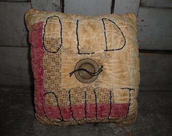 May include: A beige and red quilted pillow with the word "OLD" stitched in black thread. The pillow has a brown button in the center.
