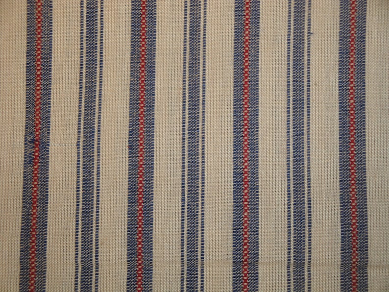 May include: Close-up of a woven fabric with vertical stripes. Alternating stripes of navy blue and red are set against a cream-coloured background. The texture of the fabric is visible, suggesting a natural fibre.