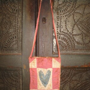 Op de afbeelding: Een kleine, handgemaakte roze en cr&egrave;me patchwork tas met een blauw hart applicatie. De tas heeft een lange, dunne riem en hangt tegen een donkere, getextureerde achtergrond. De tas lijkt vintage of antiek te zijn.