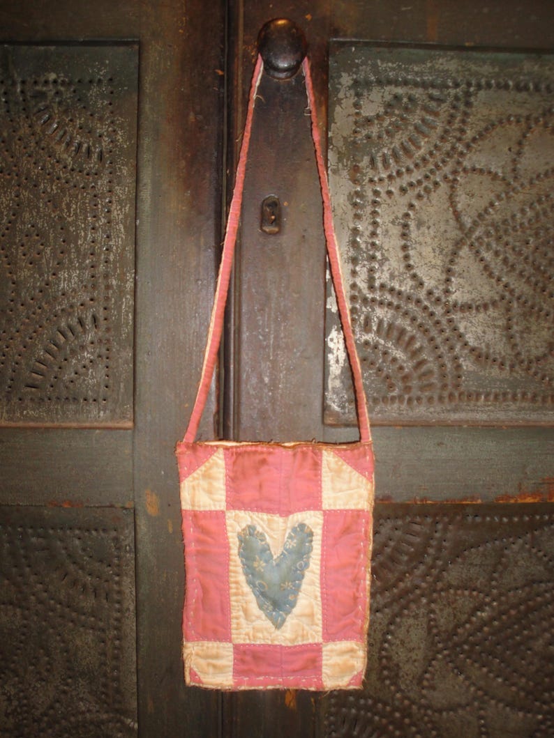 Op de afbeelding: Een kleine, handgemaakte roze en cr&egrave;me patchwork tas met een blauw hart applicatie. De tas heeft een lange, dunne riem en hangt tegen een donkere, getextureerde achtergrond. De tas lijkt vintage of antiek te zijn.
