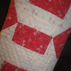 Op de afbeelding: Een doorgestikt stuk stof met een geometrisch patroon van afwisselende rode en witte rechthoekige blokken. De rode secties tonen een delicate witte bloemen- en ankerprint, terwijl de witte secties een subtiel rood ankerpatroon bevatten.