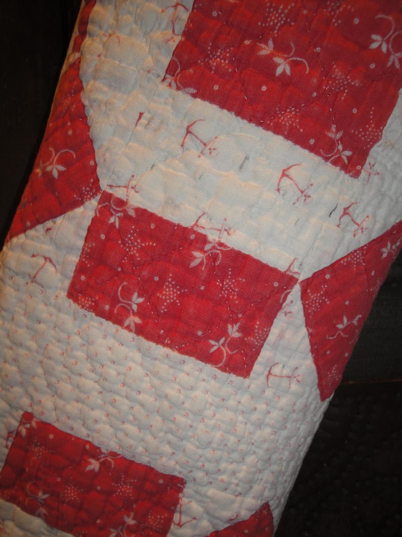 Op de afbeelding: Een doorgestikt stuk stof met een geometrisch patroon van afwisselende rode en witte rechthoekige blokken. De rode secties tonen een delicate witte bloemen- en ankerprint, terwijl de witte secties een subtiel rood ankerpatroon bevatten.