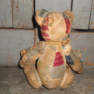 Puede incluir: Un oso de peluche hecho de varios parches de tela en tonos apagados de beige, tostado y marr&oacute;n, con detalles de patrones de cuadros y rayas en rojo, azul y verde. El oso se muestra desde atr&aacute;s, mostrando costuras visibles.