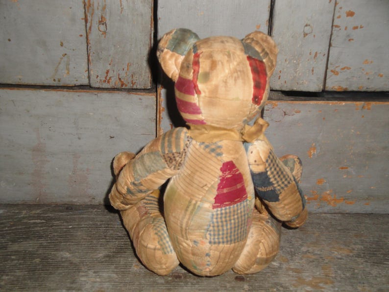 Puede incluir: Un oso de peluche hecho de varios parches de tela en tonos apagados de beige, tostado y marr&oacute;n, con detalles de patrones de cuadros y rayas en rojo, azul y verde. El oso se muestra desde atr&aacute;s, mostrando costuras visibles.