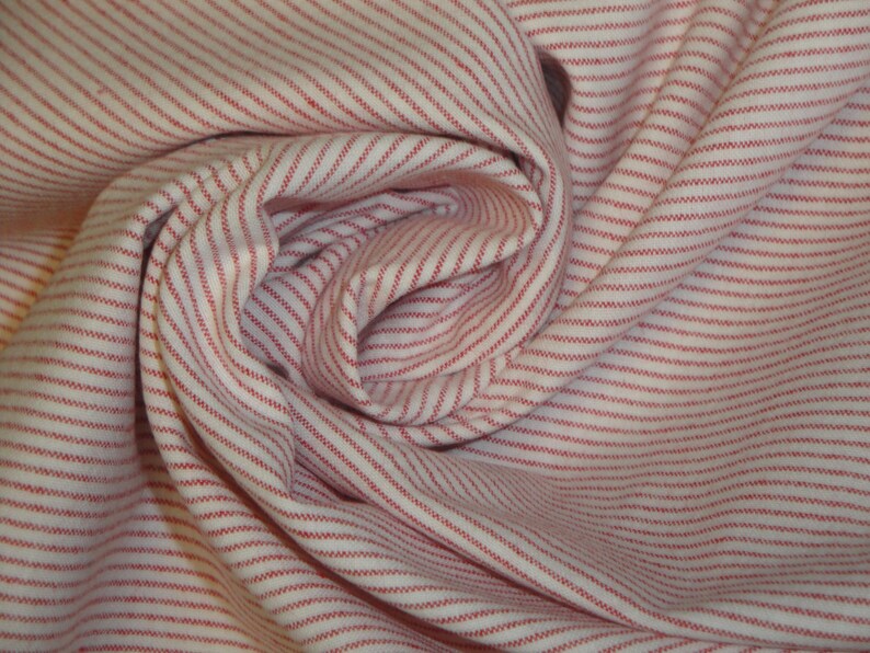 May include: Close-up of a white fabric with thin, evenly spaced red stripes. The fabric is gathered and folded, creating a swirling pattern. The stripes run parallel to each other, adding texture and visual interest to the textile.