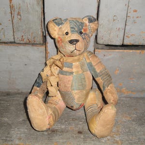 Puede incluir: Un oso de peluche construido con varios parches de tela en tonos apagados de azul, bronceado y rojo descolorido. El oso presenta ojos de bot&oacute;n oscuros, una nariz y boca cosidas en negro, y una cinta de tela deshilachada atada al cuello.