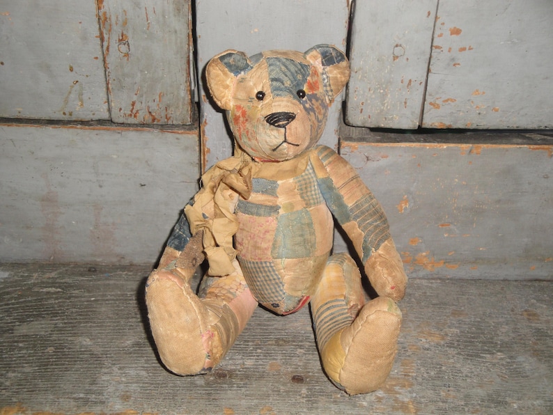 Puede incluir: Un oso de peluche construido con varios parches de tela en tonos apagados de azul, bronceado y rojo descolorido. El oso presenta ojos de bot&oacute;n oscuros, una nariz y boca cosidas en negro, y una cinta de tela deshilachada atada al cuello.