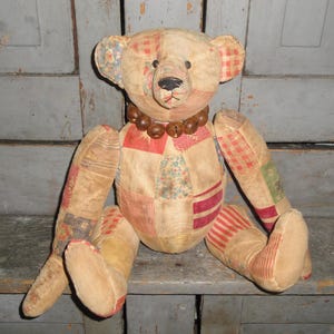 May include: A vintage patchwork teddy bear with a brown wooden bead necklace. The bear is sitting on a wooden shelf.