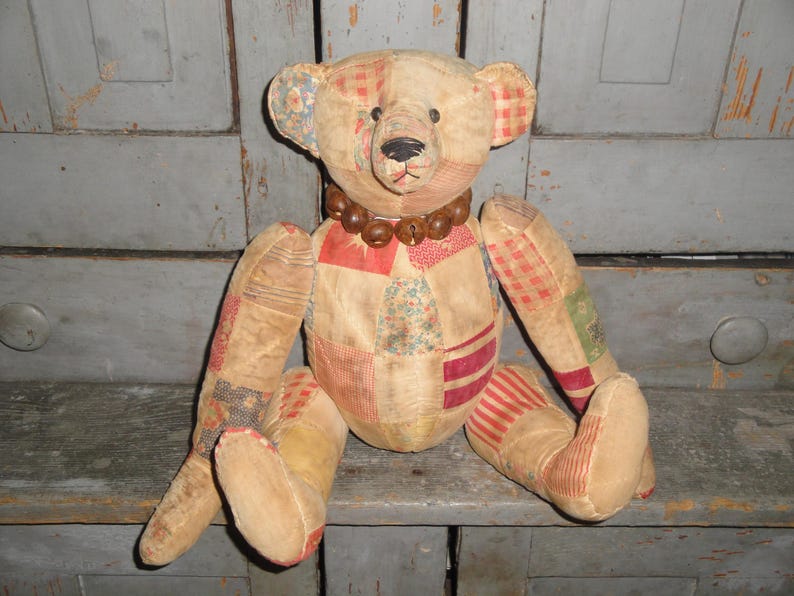 May include: A vintage patchwork teddy bear with a brown wooden bead necklace. The bear is sitting on a wooden shelf.