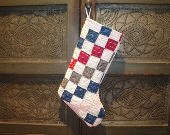 May include: A patchwork Christmas stocking with red, blue, white and grey squares. The stocking is hanging on a rustic wooden door with a decorative pattern.
