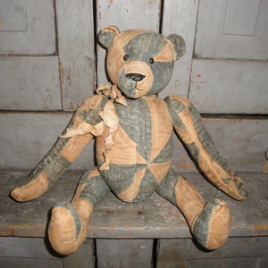 May include: A patchwork teddy bear with blue and tan fabric. The bear is sitting on a wooden shelf.