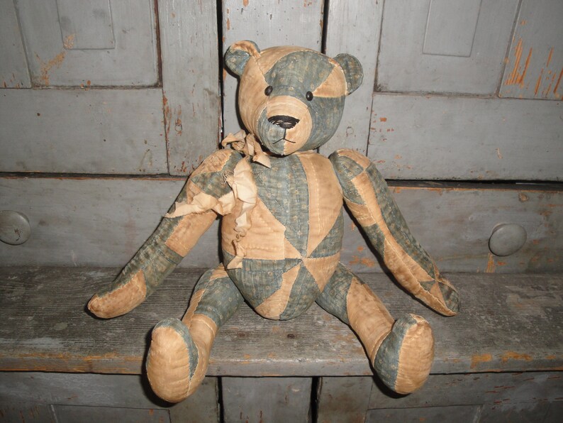 May include: A patchwork teddy bear with blue and tan fabric. The bear is sitting on a wooden shelf.