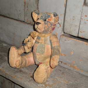 Puede incluir: Un oso de peluche construido con varios parches de tela en tonos apagados de marr&oacute;n, bronceado, azul y rojo. El oso presenta una nariz cosida, ojos oscuros y una cinta de tela deshilachada atada alrededor del cuello, con costuras visibles.