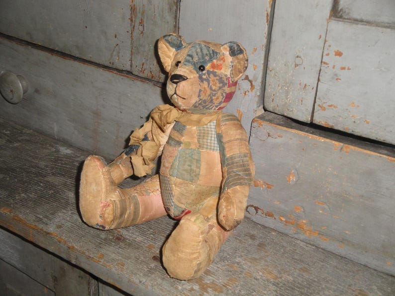 Puede incluir: Un oso de peluche construido con varios parches de tela en tonos apagados de marr&oacute;n, bronceado, azul y rojo. El oso presenta una nariz cosida, ojos oscuros y una cinta de tela deshilachada atada alrededor del cuello, con costuras visibles.