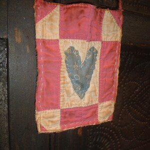 Op de afbeelding: Een kleine, handgemaakte roze en cr&egrave;mekleurige patchworktas met een blauw hartje. De tas heeft een lange, dunne riem en is gemaakt van gewatteerde stof. Het hart is versierd met een bloemenpatroon.