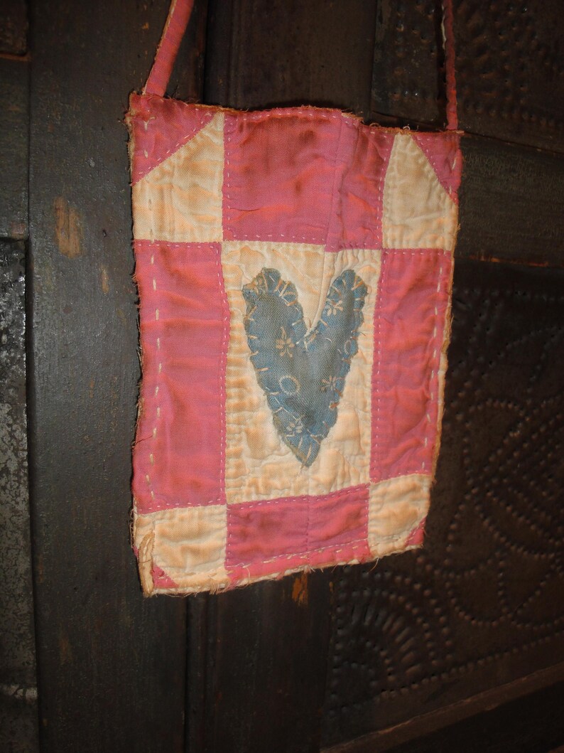 Op de afbeelding: Een kleine, handgemaakte roze en cr&egrave;mekleurige patchworktas met een blauw hartje. De tas heeft een lange, dunne riem en is gemaakt van gewatteerde stof. Het hart is versierd met een bloemenpatroon.