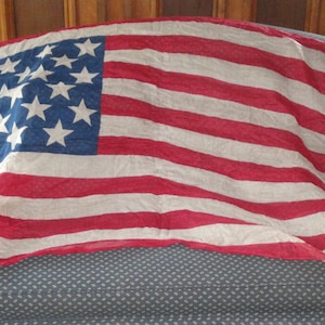 Op de afbeelding: Een vintage Amerikaanse vlag met 13 rode en witte strepen en 20 witte sterren op een blauw veld. De vlag is gemaakt van een dunne, verbleekte stof en is gedrapeerd over een blauwe bank.