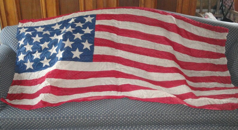 Op de afbeelding: Een vintage Amerikaanse vlag met 13 rode en witte strepen en 20 witte sterren op een blauw veld. De vlag is gemaakt van een dunne, verbleekte stof en is gedrapeerd over een blauwe bank.