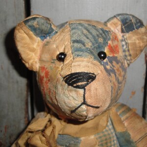 Puede incluir: Un oso de peluche construido con varios parches de tela en tonos bronceado, azul y patrones florales apagados. El oso presenta dos peque&ntilde;os ojos de cuentas negras y una nariz y boca cosidas a mano con hilo negro, con costuras visibles.