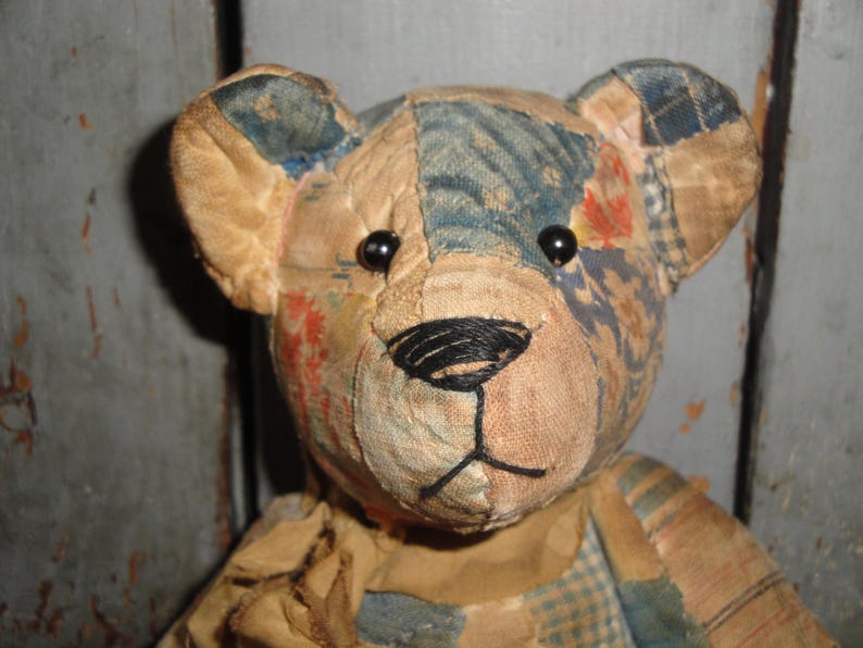 Puede incluir: Un oso de peluche construido con varios parches de tela en tonos bronceado, azul y patrones florales apagados. El oso presenta dos peque&ntilde;os ojos de cuentas negras y una nariz y boca cosidas a mano con hilo negro, con costuras visibles.