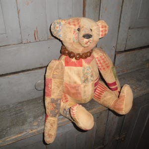 May include: A patchwork teddy bear with a brown bead necklace. The bear is sitting on a wooden surface.