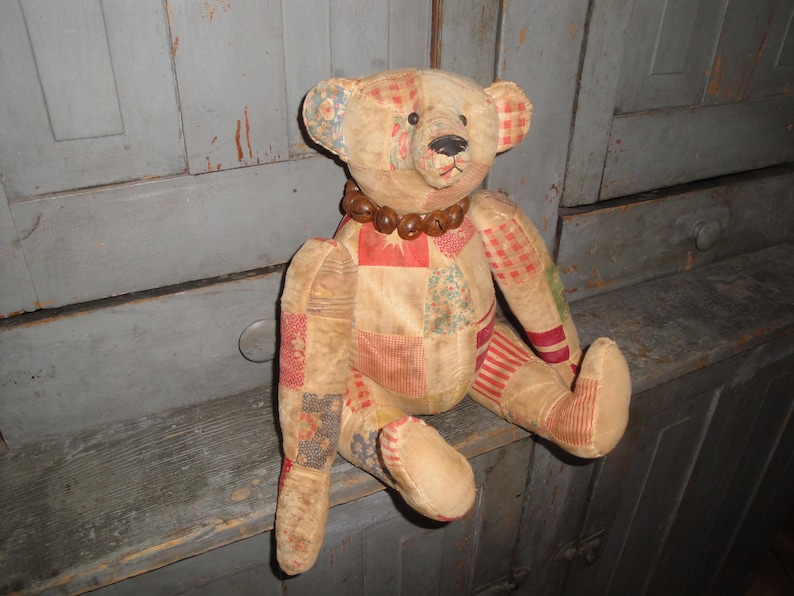 May include: A patchwork teddy bear with a brown bead necklace. The bear is sitting on a wooden surface.