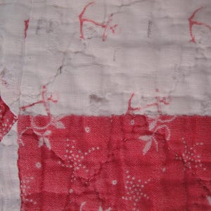 Op de afbeelding: Een gewatteerd textiel met een patchworkontwerp met afwisselende witte en rode stoffen vierkanten. De witte secties tonen een vaag rood bloemenpatroon, terwijl de rode secties een witte bloemenprint hebben. De stof vertoont zichtbare stiksels.