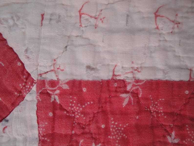 Op de afbeelding: Een gewatteerd textiel met een patchworkontwerp met afwisselende witte en rode stoffen vierkanten. De witte secties tonen een vaag rood bloemenpatroon, terwijl de rode secties een witte bloemenprint hebben. De stof vertoont zichtbare stiksels.