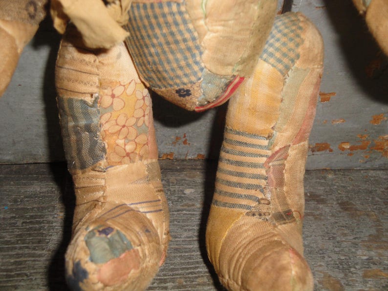 Puede incluir: Una figura de juguete rellena construida con varios parches de tela en tonos beige, bronceado y crema. Las piezas textiles presentan diversos patrones, incluyendo cuadros vichy azules y blancos, estampados florales y rayas finas.