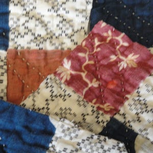 May include: Close-up of a patchwork quilt with a variety of fabrics and patterns. The quilt features squares of indigo blue, white with black geometric designs, rust-colored, and burgundy with floral patterns. The stitching is visible.
